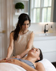 A person receiving a facial treatment lying on a table with a aesthetically pleasing room setting, including a seated female aesthetician attending to the client.