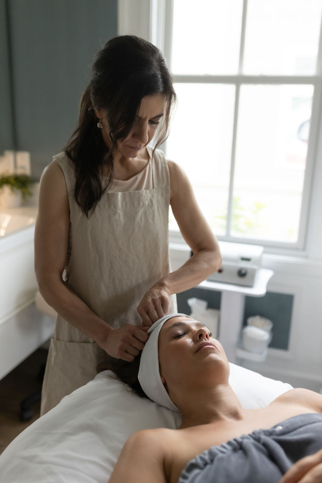 A person receiving a facial treatment, lying on a treatment bed with a focus on a facial mask being applied by a seated person in a spa setting.
