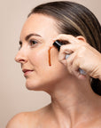 Woman applying a brown liquid to her face with a brush against a beige background