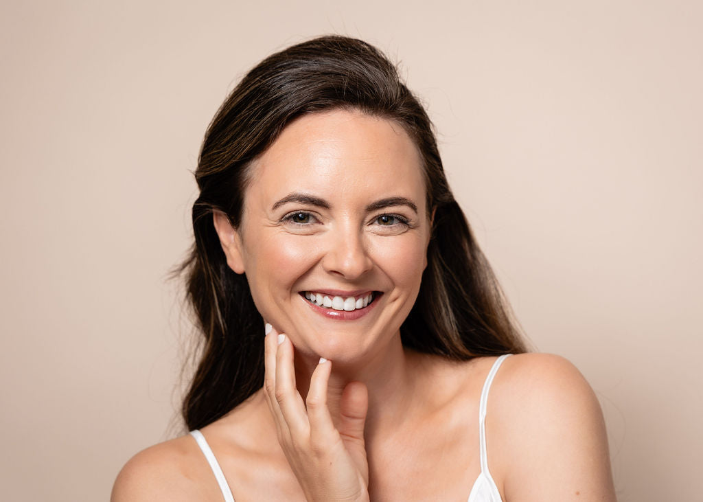 Woman smiling and touching her face against a beige background with Geri G. products