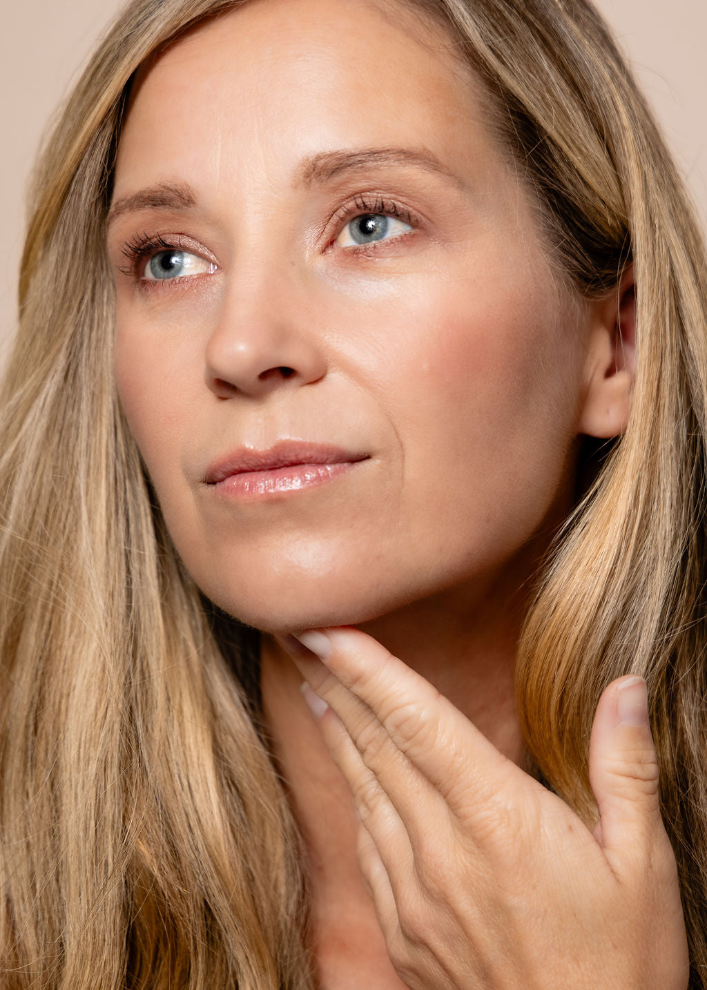 Close-up of a woman with long blonde hair touching her face against a neutral background